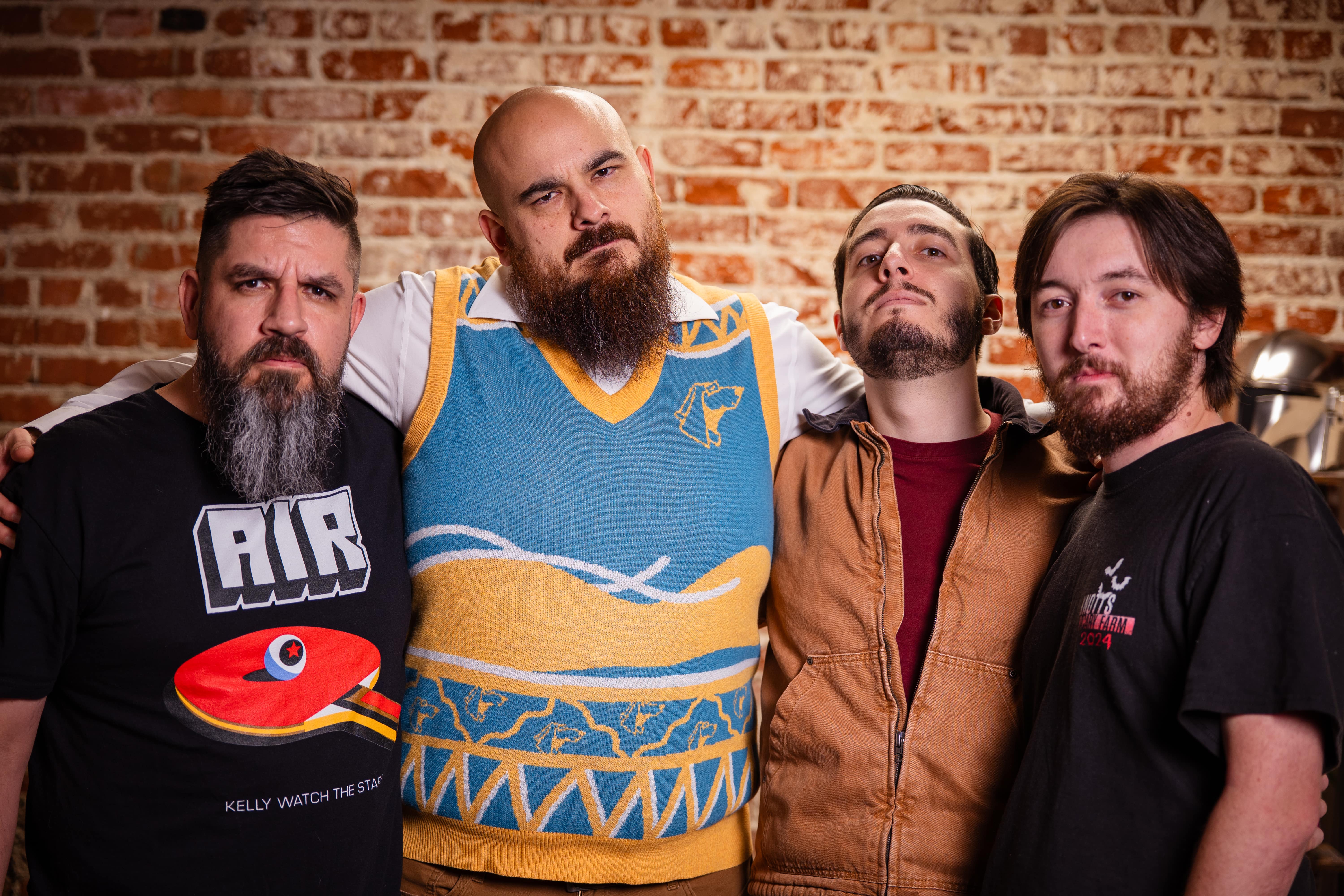 Four adult men stand shoulder to shoulder in a casual group portrait against a brick wall, posing with relaxed but serious expressions.