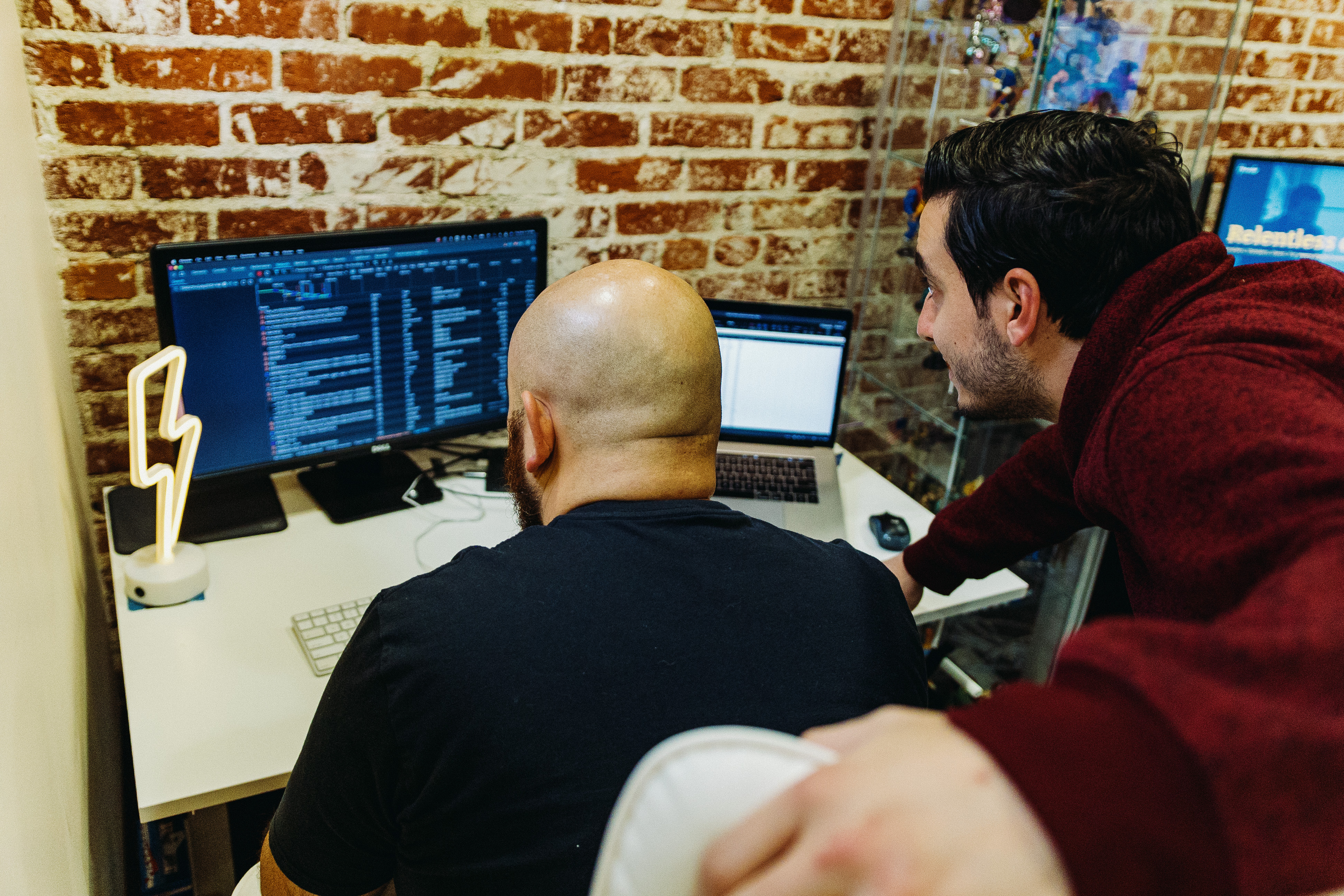 Two men working on computer screens in a brick-walled office.