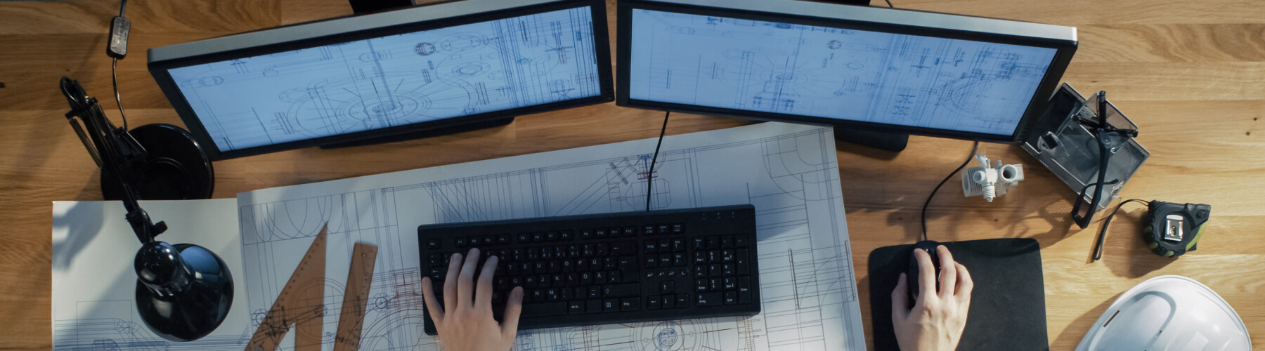 Engineer working at dual monitors with blueprints on desk.