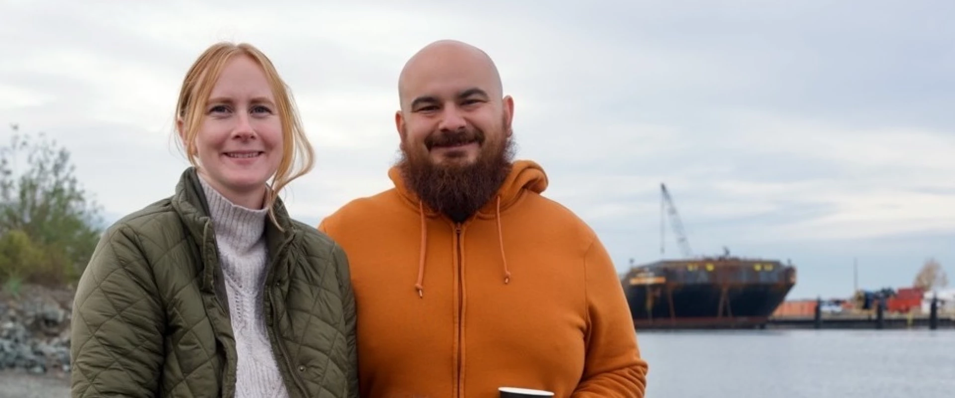 Smiling couple poses by waterfront with ship in the background on a cloudy day.