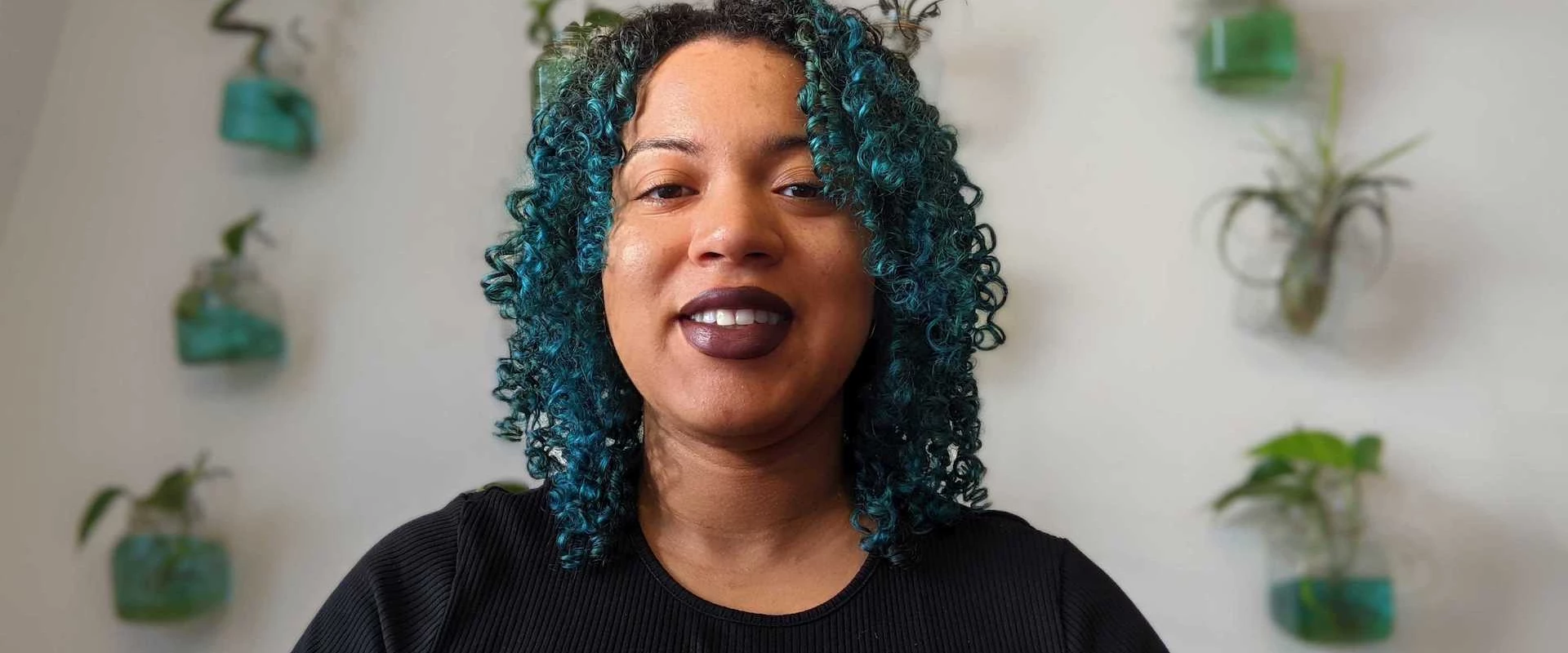 Person with curly teal hair and dark lipstick smiling indoors.