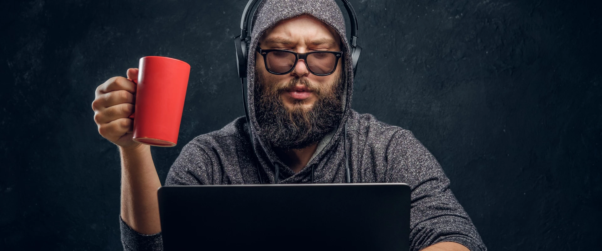 Man in hoodie and headphones holds red mug while working on laptop.