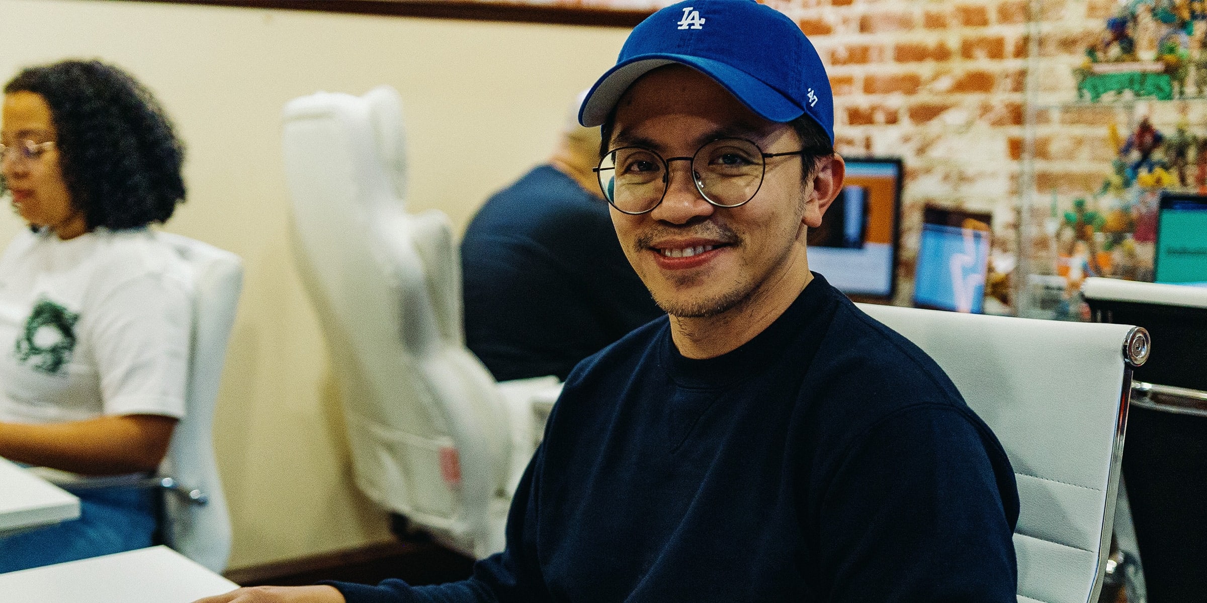 Man in blue cap and glasses smiling at desk in office setting.