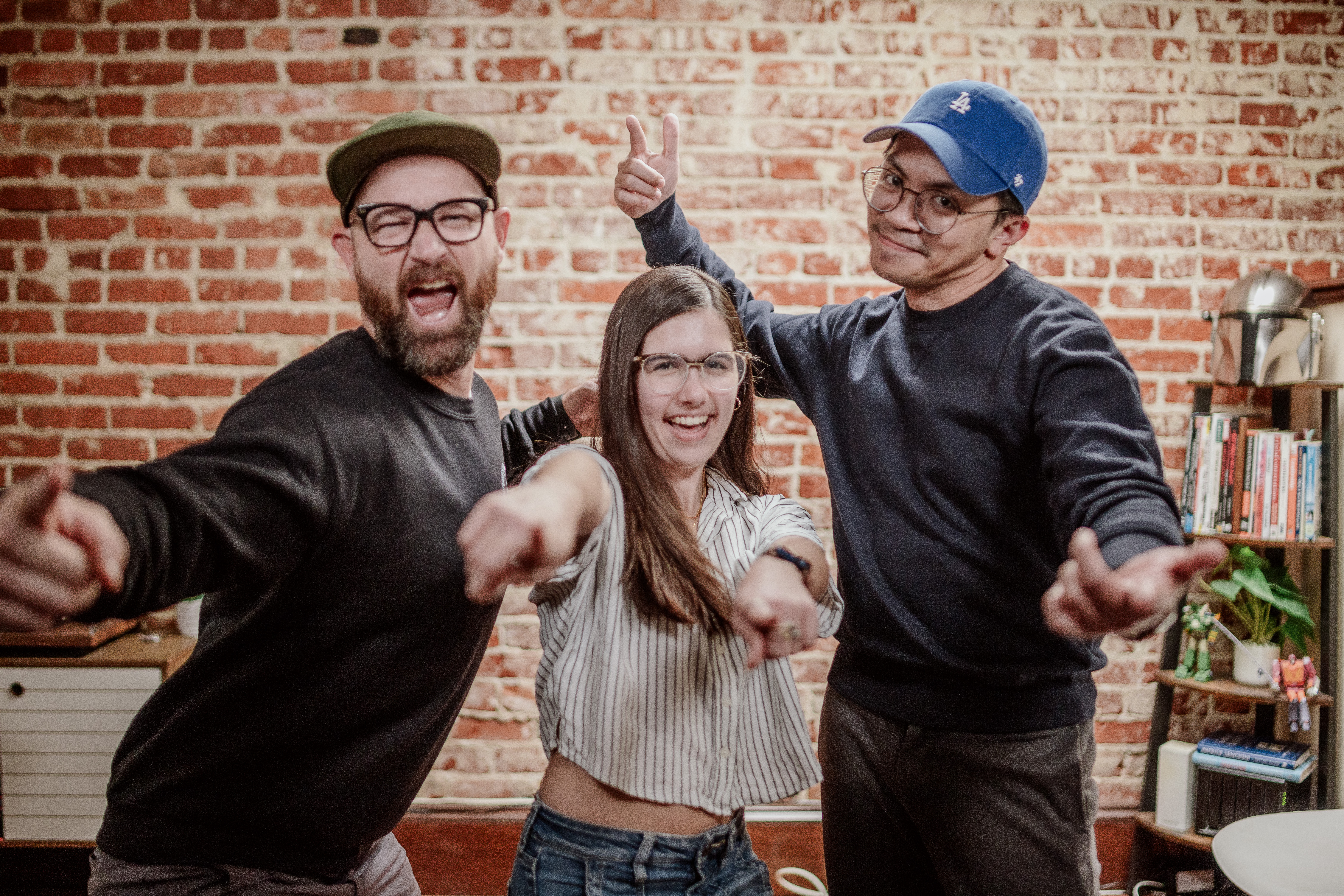 Three friends smiling and pointing at the camera in front of a brick wall.