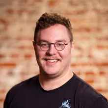 Smiling person with glasses against a blurred brick background.