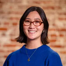 Smiling person with glasses, wearing a blue sweater, brick background.