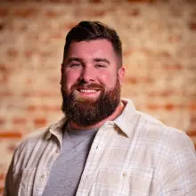 Bearded man in plaid shirt smiling against brick wall.