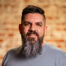 Smiling man with a long beard, blurred brick wall background.
