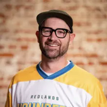 Man smiling in a yellow and white sports jersey, wearing glasses and a green cap.