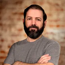 Man with a beard and gray t-shirt standing against a blurred brick background.