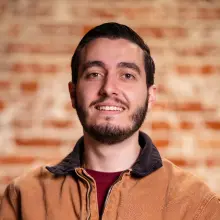 Smiling man in a brown jacket against a brick wall backdrop.