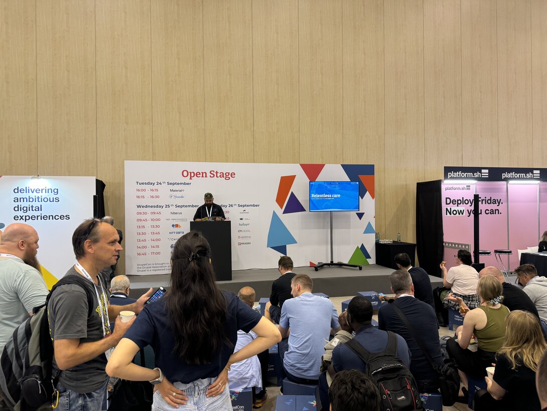 People attend a tech conference presentation at an open stage with digital experience displays.