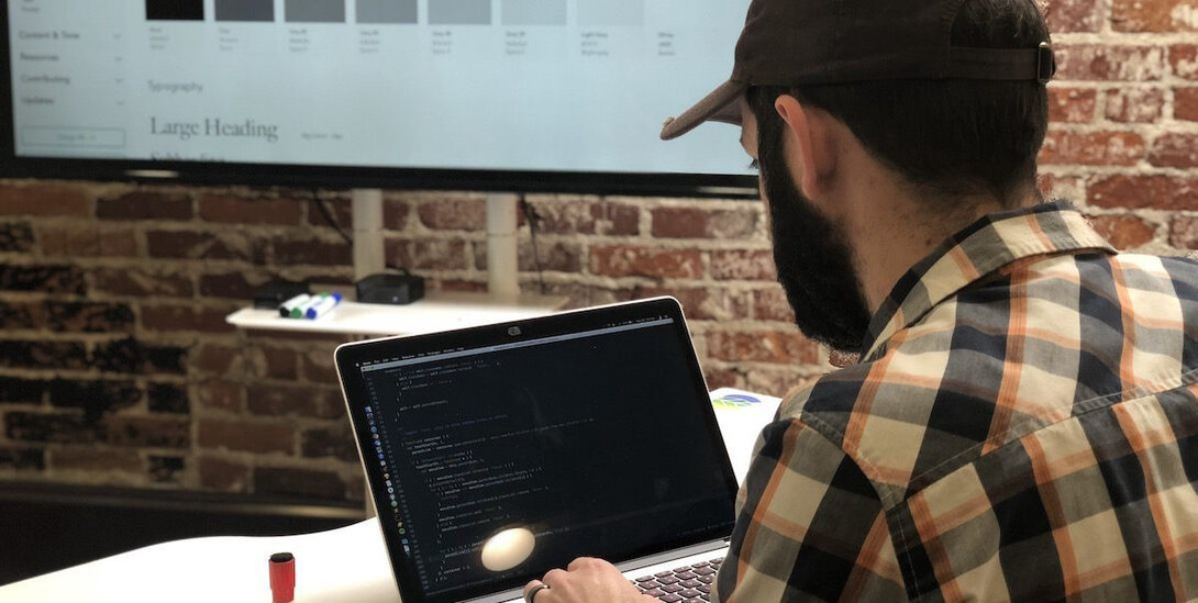 Developer coding on a laptop with a design board in the background.