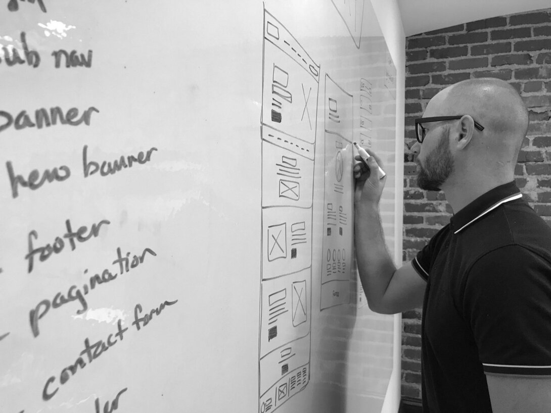 Man sketching website design on whiteboard in brick-walled office.
