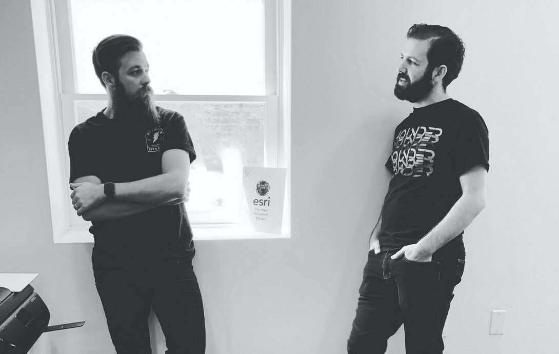 Two men with beards conversing by a window with an Esri certification visible.