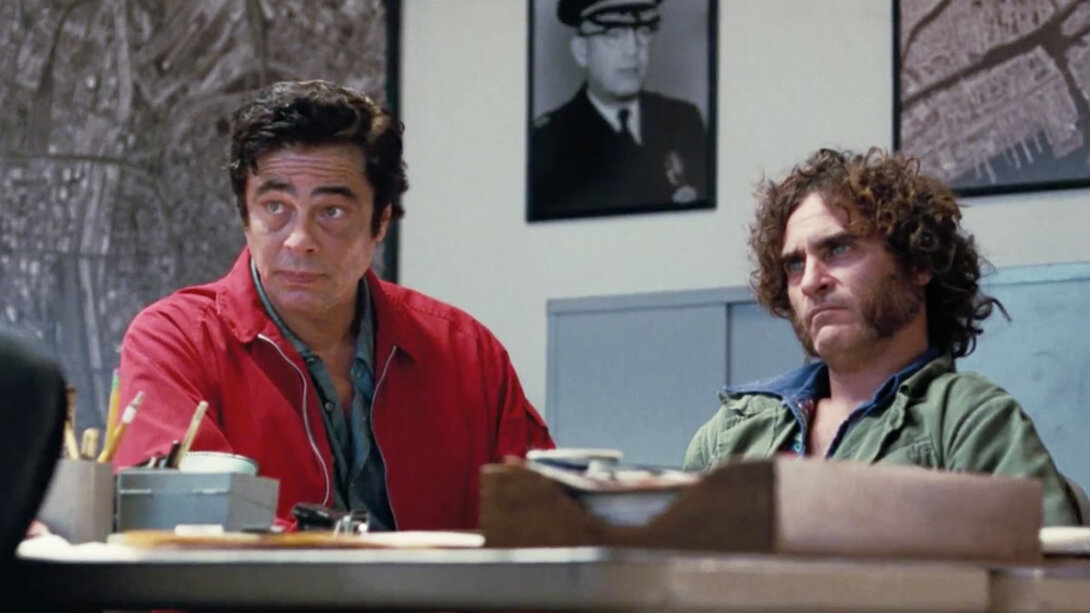 Two men sitting at a cluttered table, looking serious in a room with maps and a police officer's portrait.