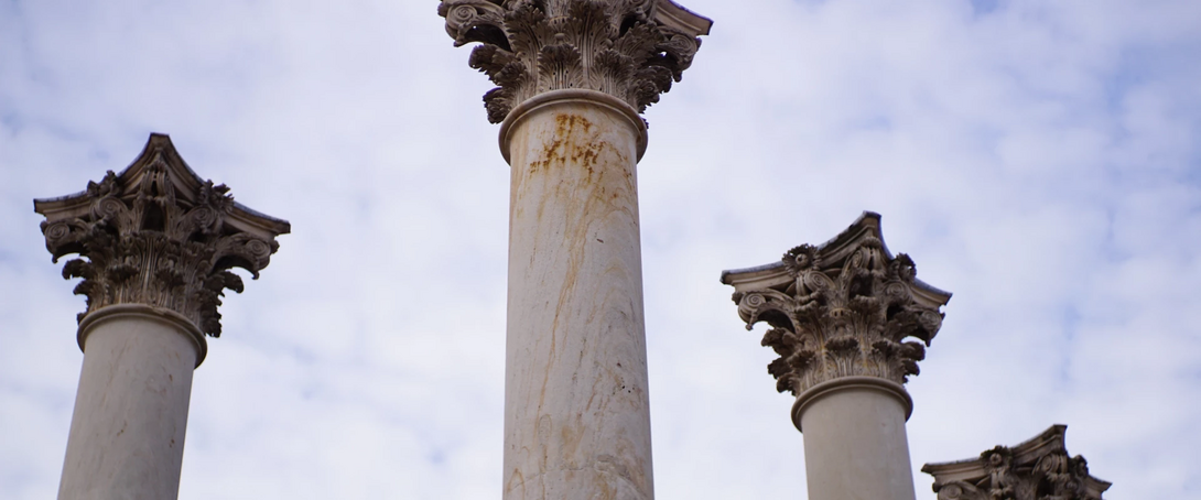 Three ornate Corinthian columns against a cloudy sky.