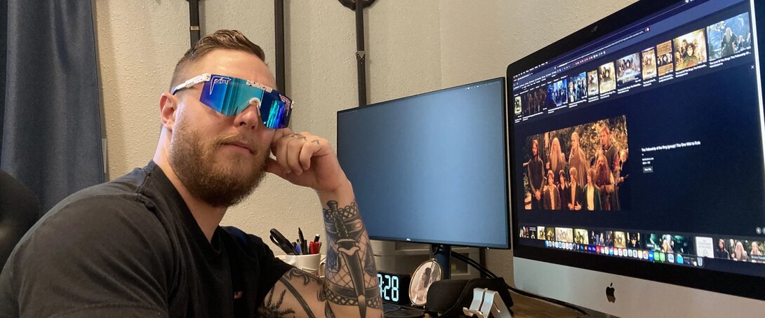 Man wearing reflective sunglasses at a desk with dual monitors displaying computer screens.
