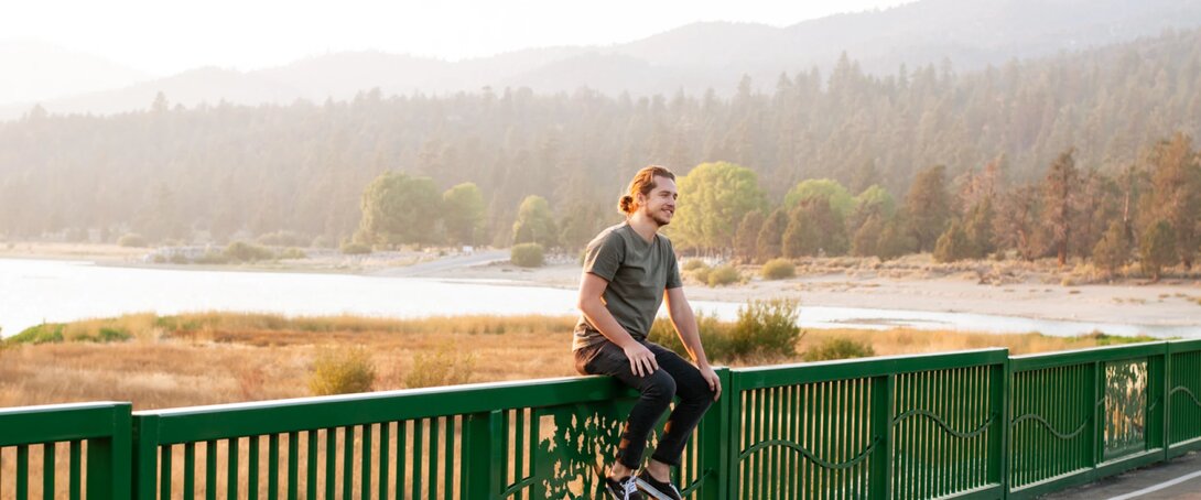 Man sitting on a green fence overlooking a serene lakeside landscape.