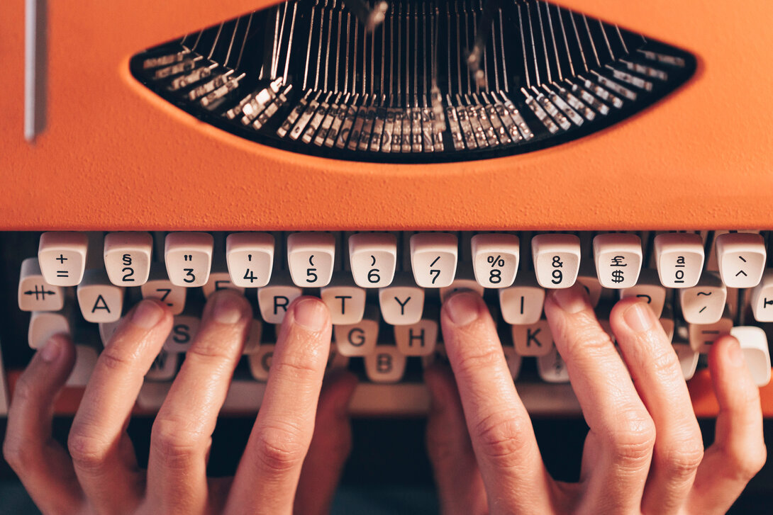 Hands typing on an orange vintage typewriter keyboard.