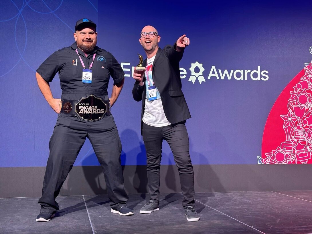 Two men celebrate on stage at the Acquia Engage Awards event, one wearing a championship belt.